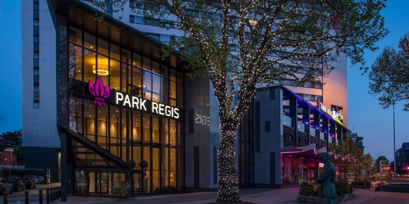 The Park Regis Birmingham exterior at dusk, its illuminated sign and a tree strung with lights beside the glass entrance